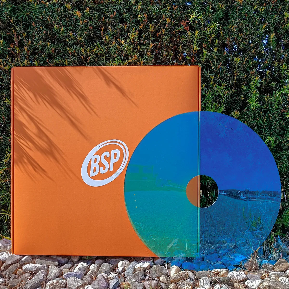 Blauer Rollstuhl-Speichenschutz nahe orangefarbener BSP-Verpackung auf grünem Gras und Steinen präsentiert