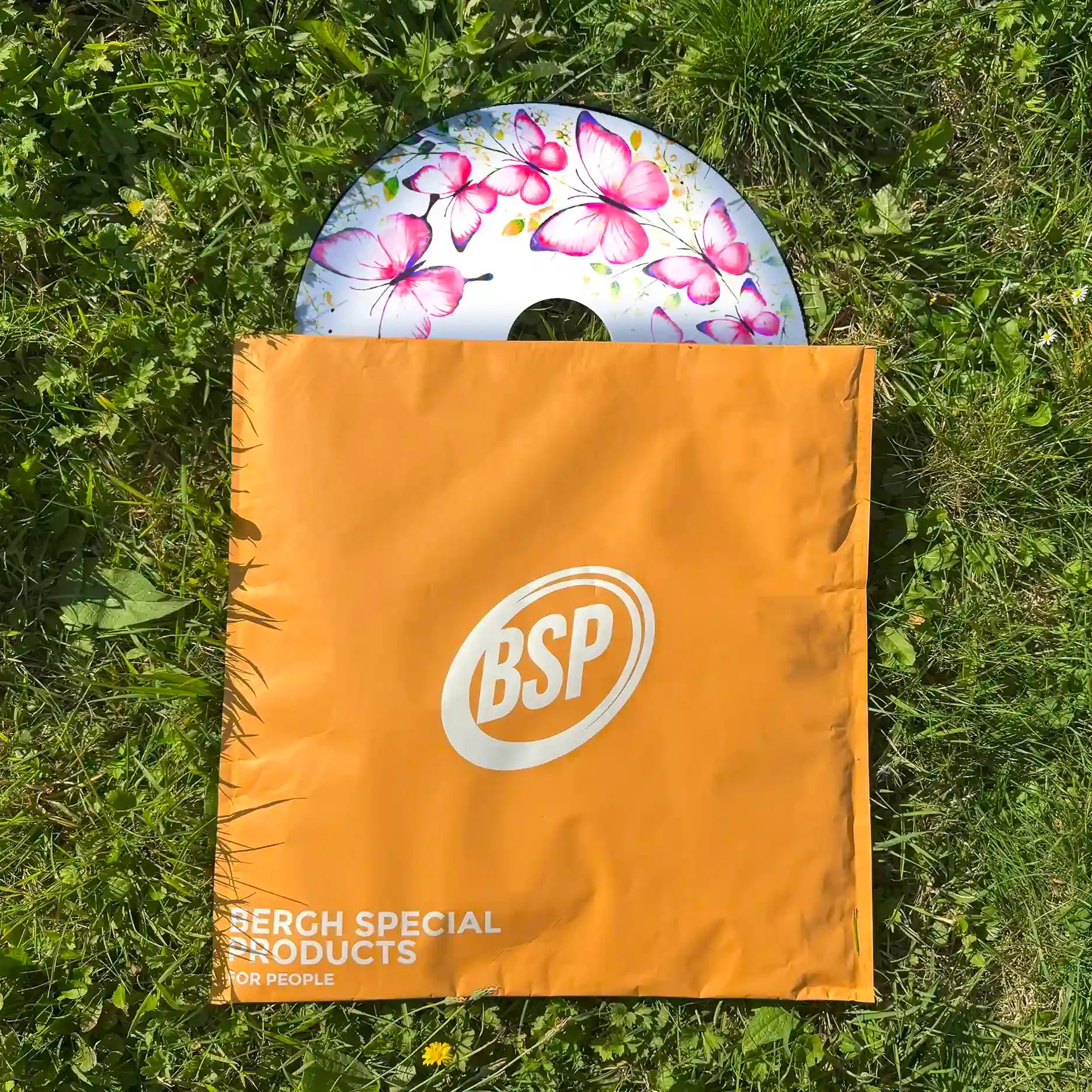Butterfly wheelchair spoke guard with pink and purple design, partially in orange BSP bag on grass.