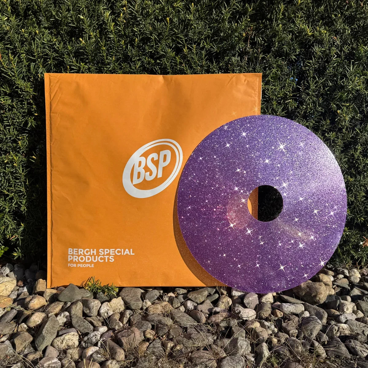 Violetter Glitzer-Rollstuhl-Speichenschutz mit Sternenmuster neben orangefarbener BSP-Verpackung auf Kies