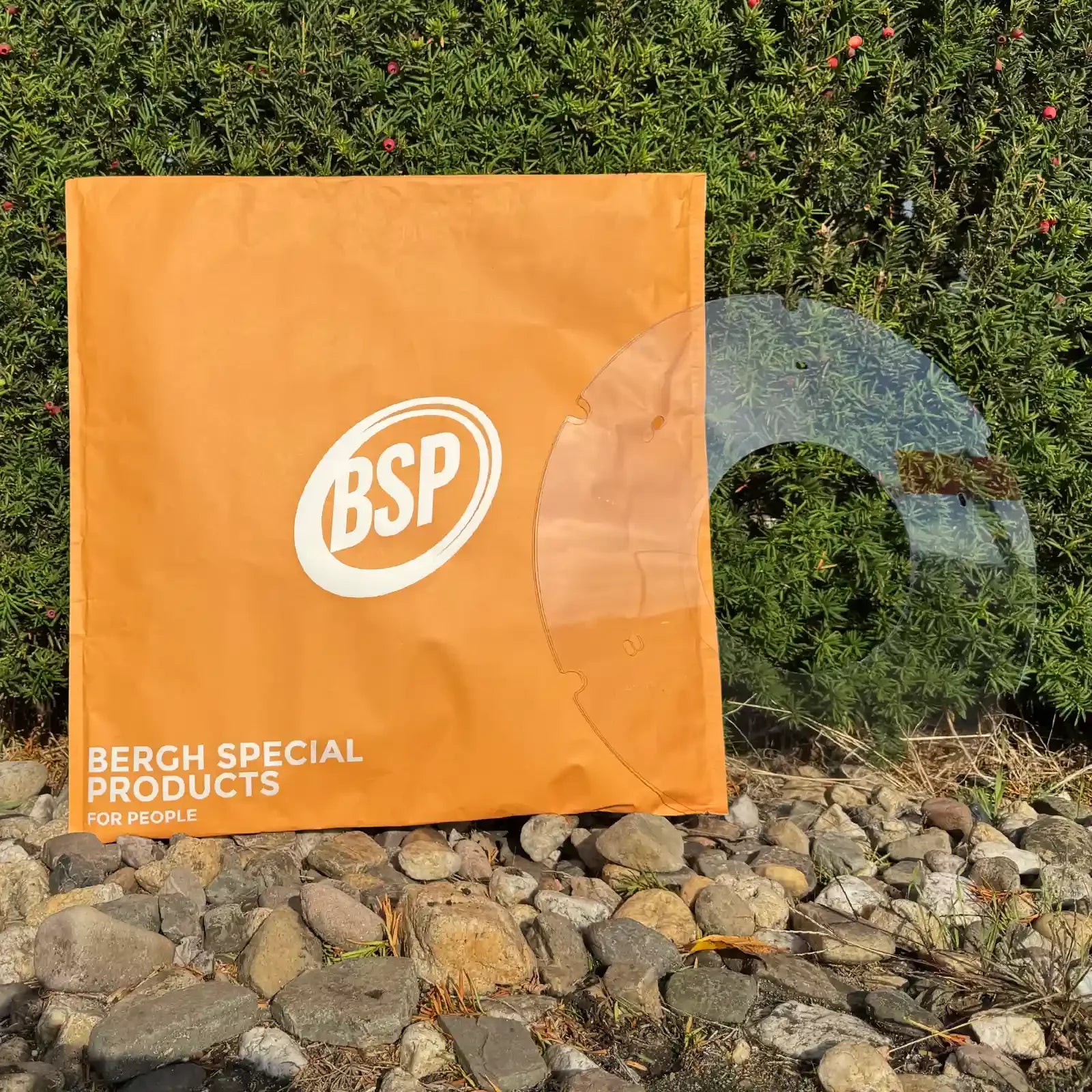 Transparent wheelchair spoke guard displayed outdoors near a green hedge and orange BSP bag.