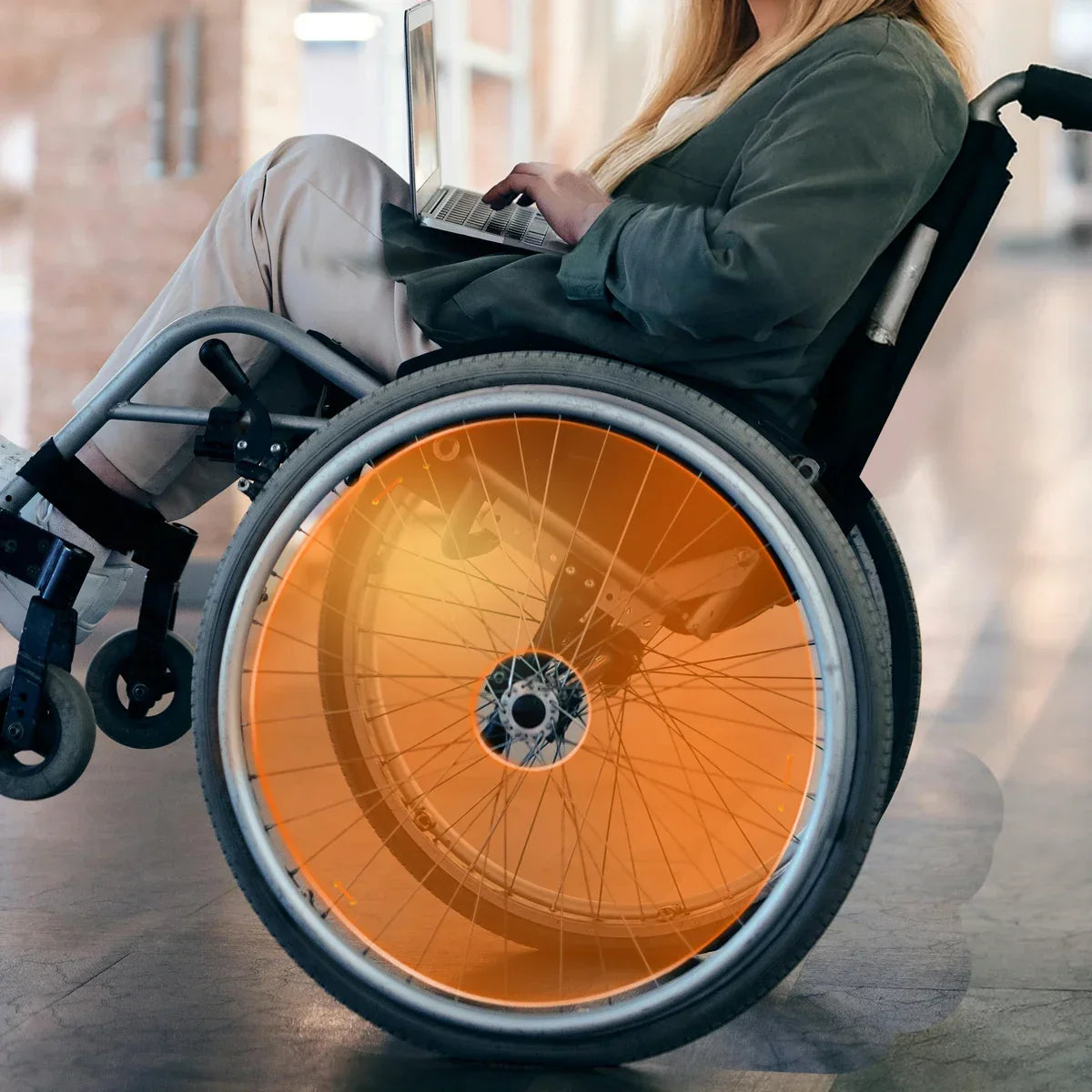Woman using a wheelchair with bright orange spoke guards indoors, working on a laptop