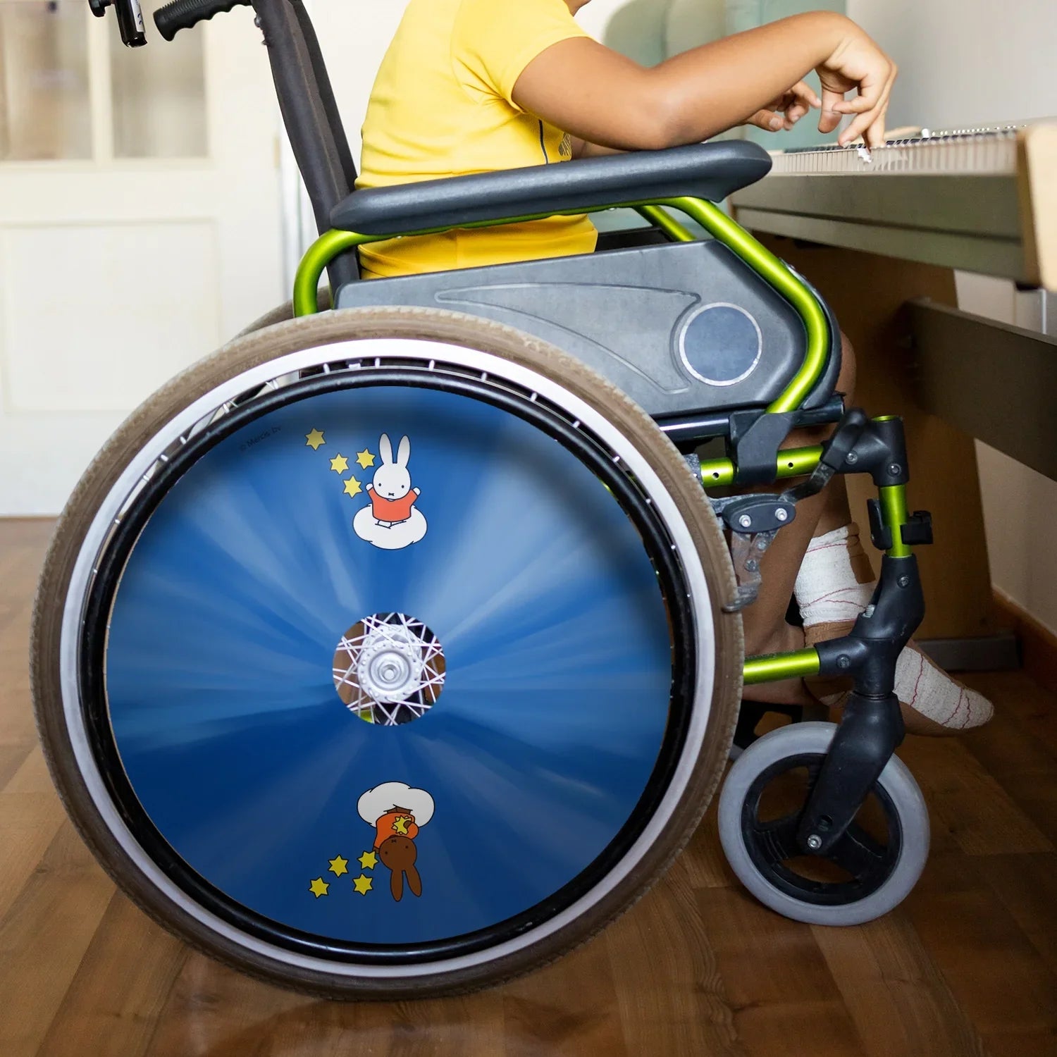 Child in wheelchair with blue Miffy and Melanie star-themed spoke guard, indoor setting
