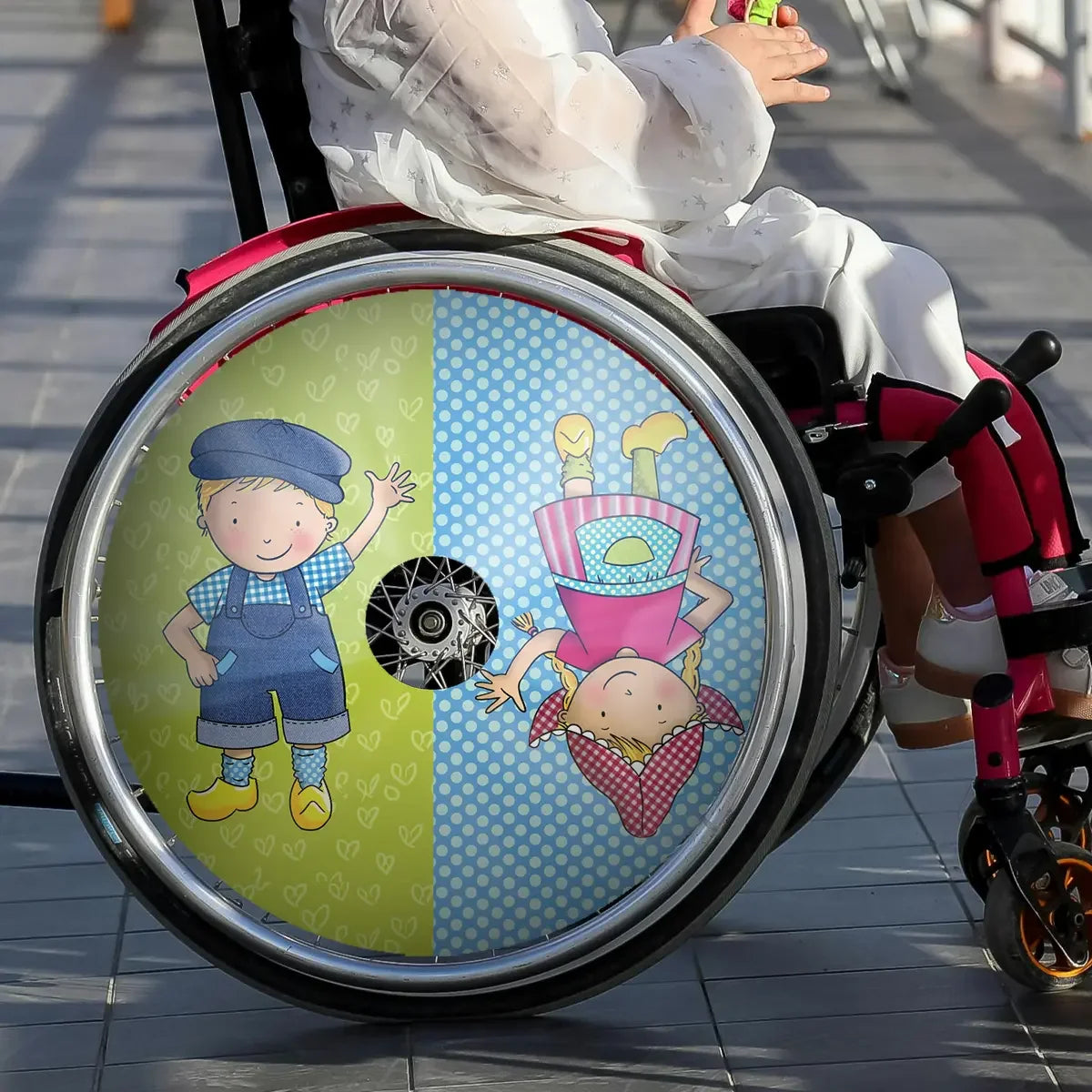Child in wheelchair with colorful spoke guard featuring cartoon boy and girl illustrations