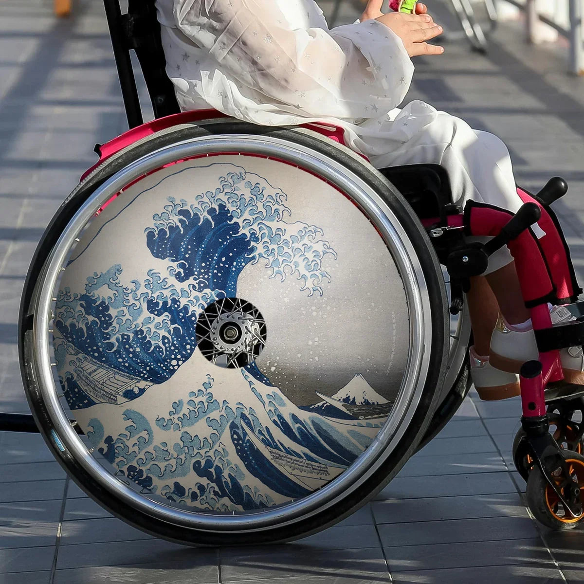 Girl using wheelchair with The Great Wave art spoke guard accessory, outdoors on tiled floor