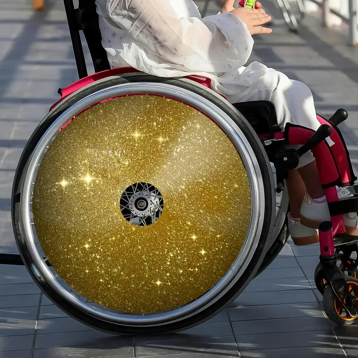 Child in wheelchair with gold glitter spoke guard, wearing white outfit, indoor setting