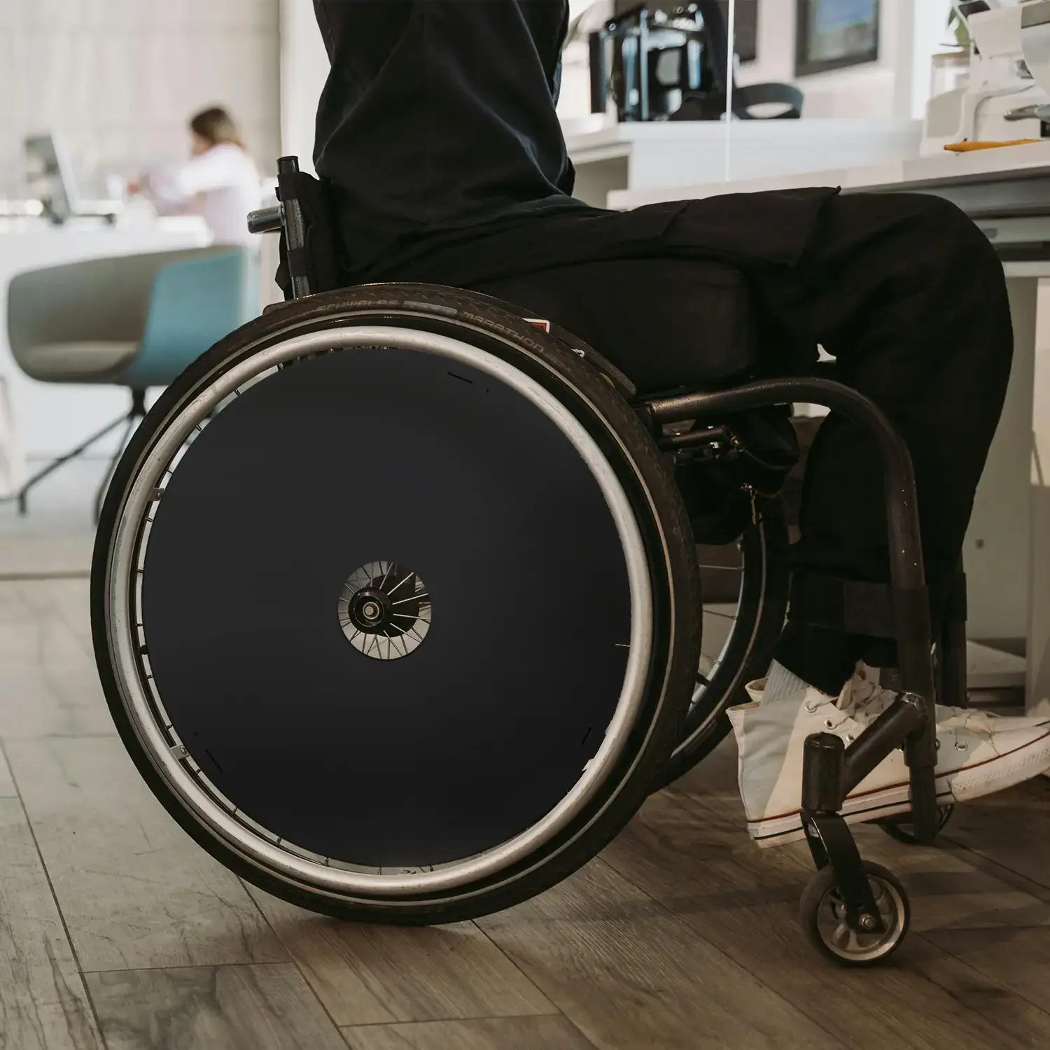 Person in wheelchair with black spoke guards, seated indoors on wood floor