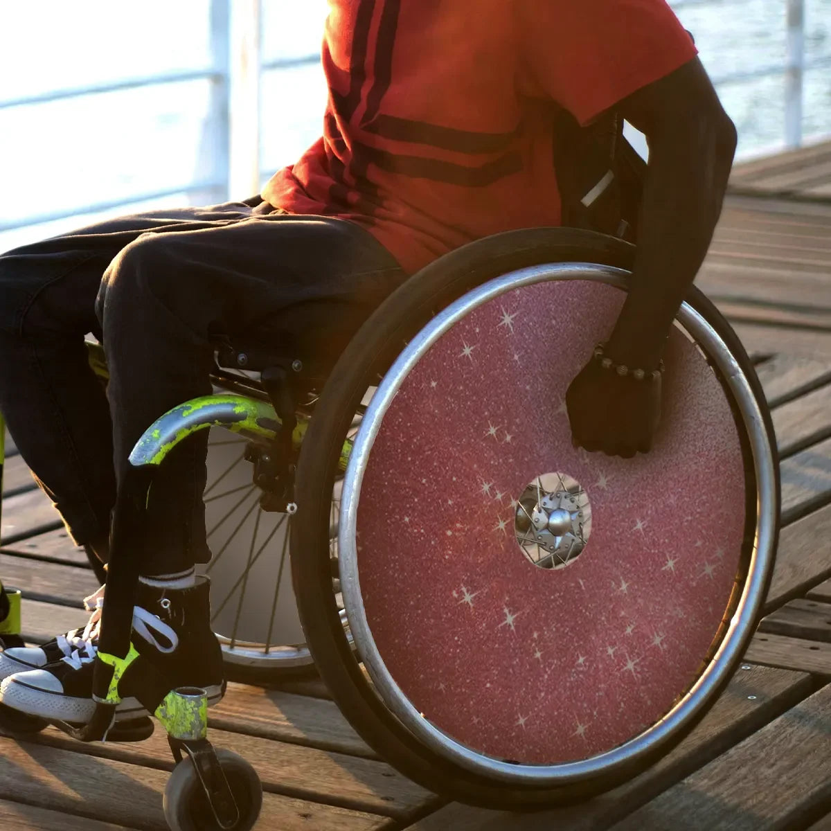 Person using wheelchair with pink starry spoke guards on wooden deck near water