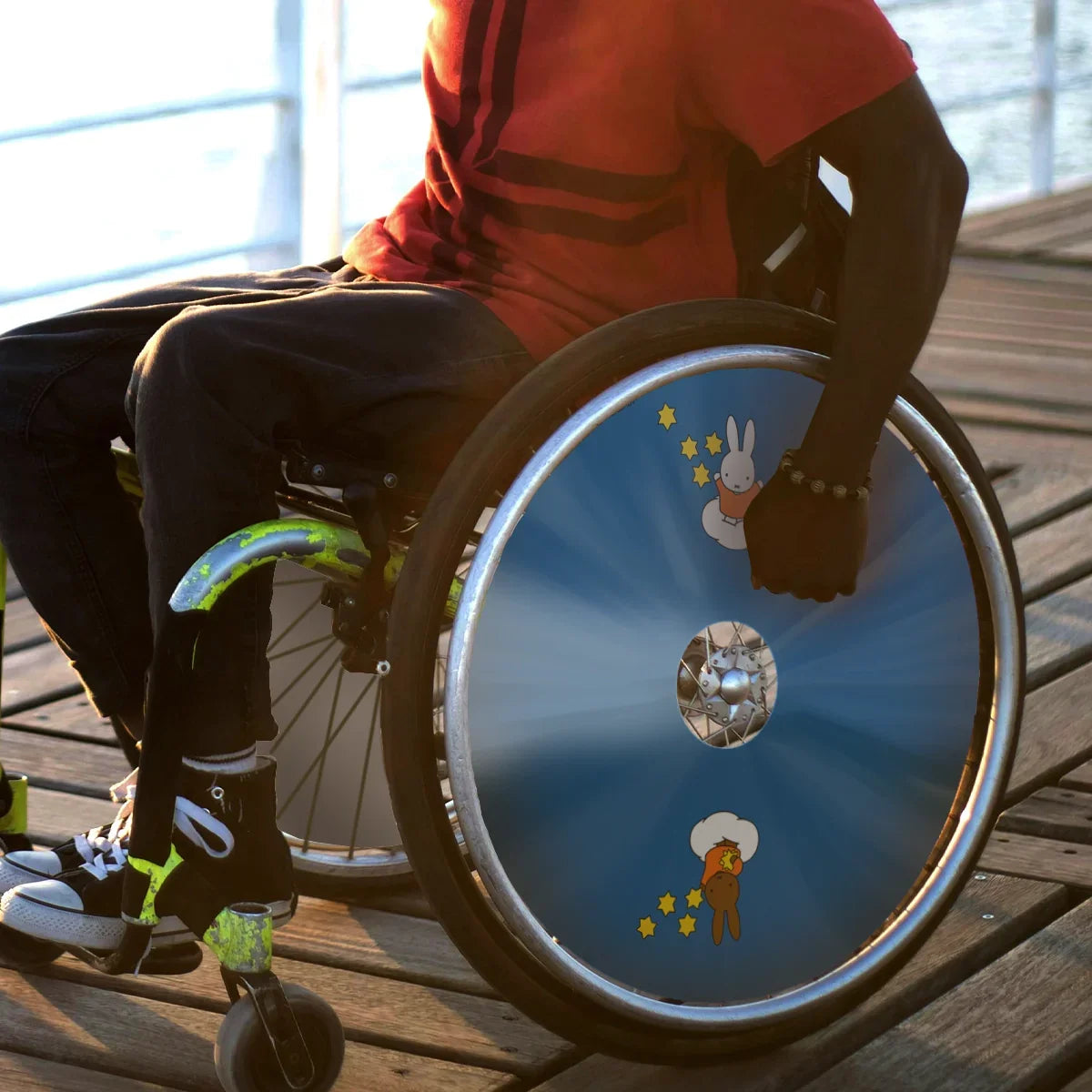 Man in wheelchair with blue spoke guards featuring cartoon rabbits and stars design on outdoor boardwalk