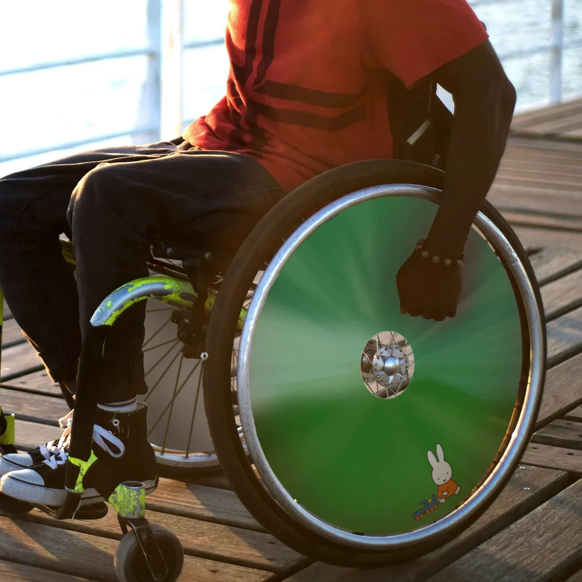 Man in wheelchair with green Miffy toy train spoke guard on wooden boardwalk