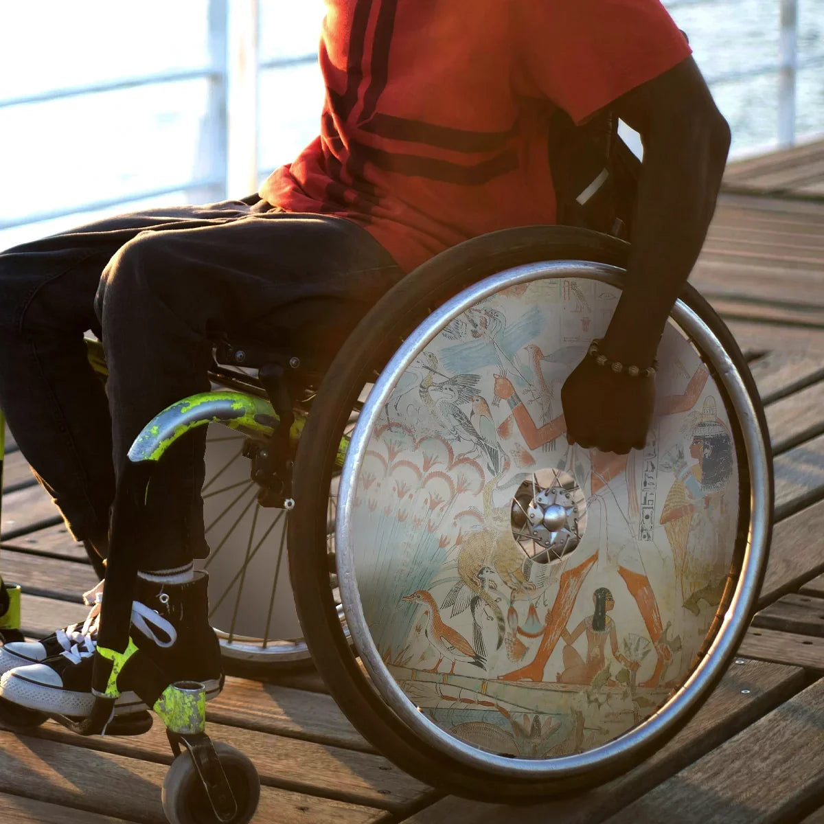 Man in wheelchair with artistic spoke guards on wheels, sitting on wooden deck outdoors