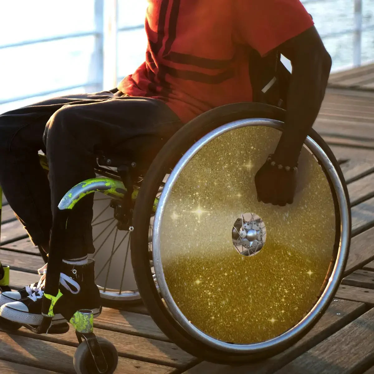 Wheelchair with gold glitter spoke guards on a wooden boardwalk, person in red shirt