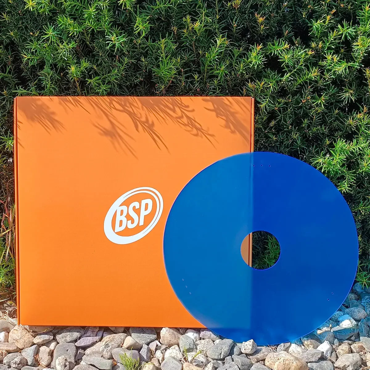Blue wheelchair spoke guard with orange BSP box on gravel, green foliage background