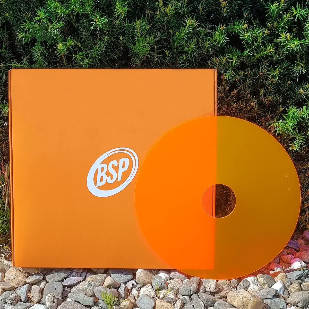 Orange BSP-branded wheelchair spoke guard with matching box, set against green foliage and pebbles