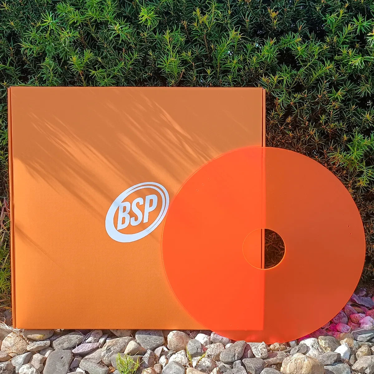 Orange BSP wheelchair spoke guard with matching box, set against green foliage and pebbles