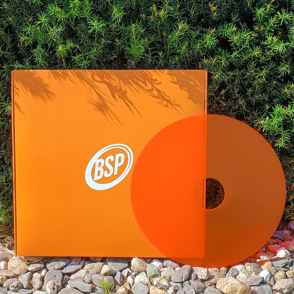Orange BSP-branded box and disc on pebbles with green foliage, showcasing wheelchair accessory packaging