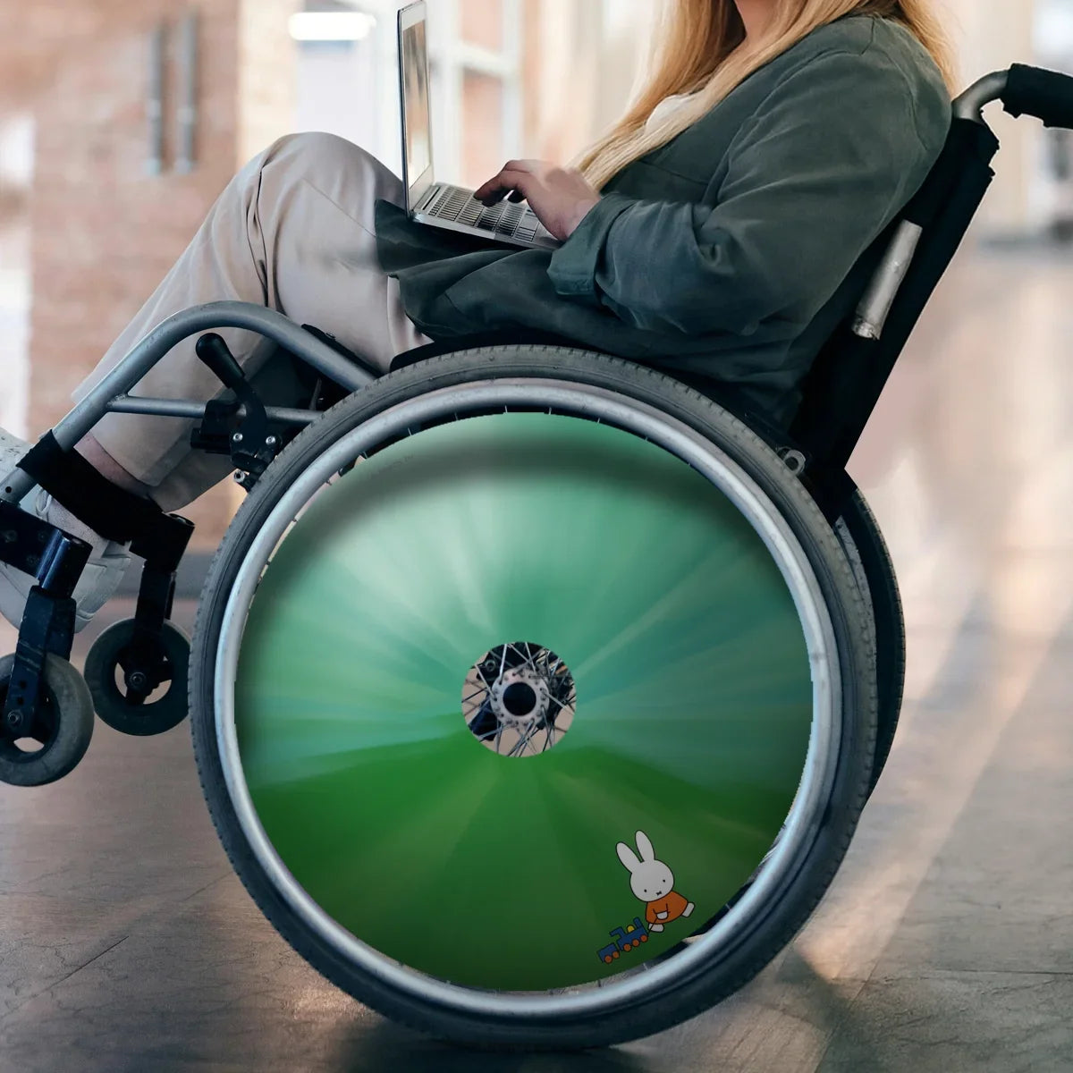 Woman in wheelchair with green Miffy-themed spoke guard, using laptop indoors