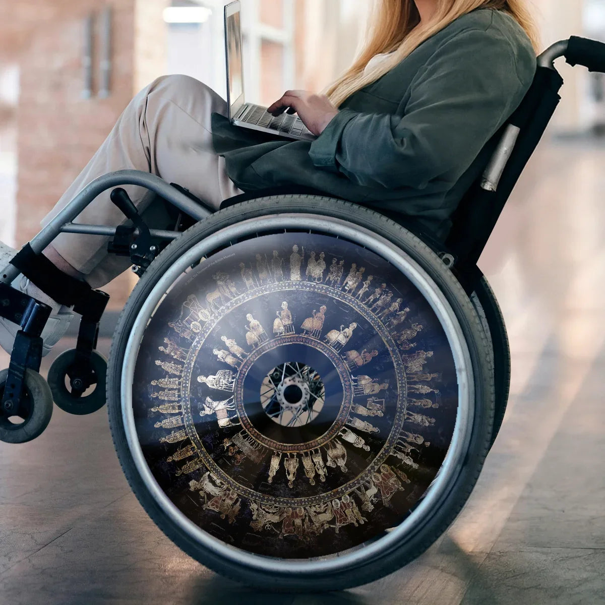 Frau im Rollstuhl mit Speichenschutz im Design der Standarte von Ur, arbeitet am Laptop im Innenbereich