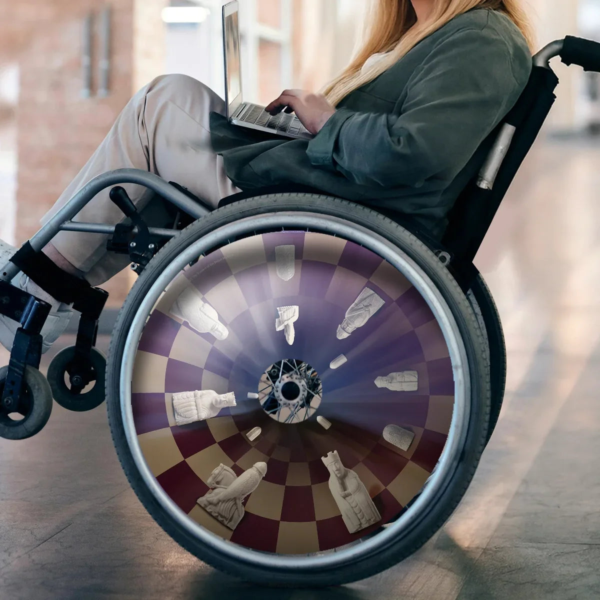 Woman in wheelchair with decorative chess-themed spoke guard, using laptop indoors